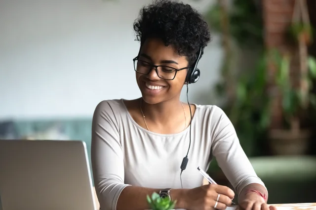 woman with headset smiling looking at laptop taking notes